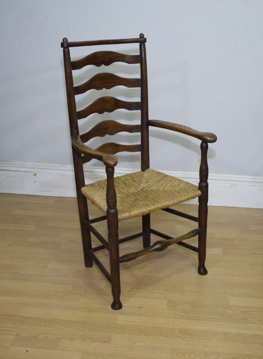 A 19th-century dark oak ladder-back armchair featuring five shaped horizontal back slats, turned legs, curved armrests, and a woven rush seat. View VI