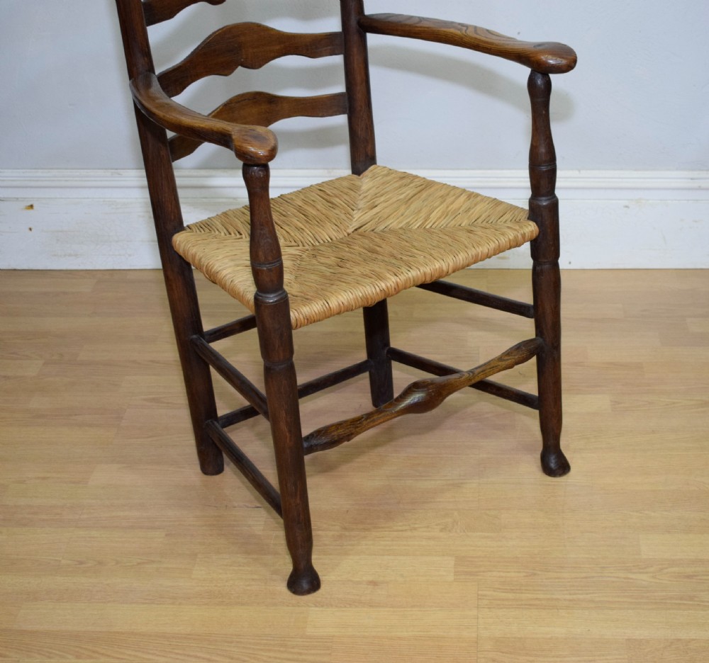 A 19th-century dark oak ladder-back armchair featuring five shaped horizontal back slats, turned legs, curved armrests, and a woven rush seat. View VII