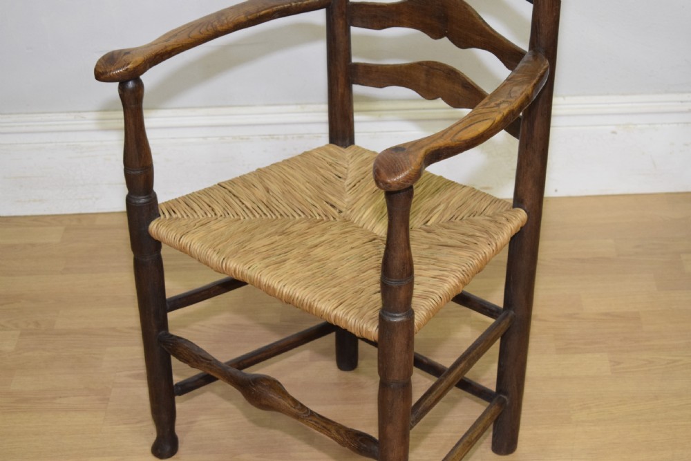 A 19th-century dark oak ladder-back armchair featuring five shaped horizontal back slats, turned legs, curved armrests, and a woven rush seat. View XIV
