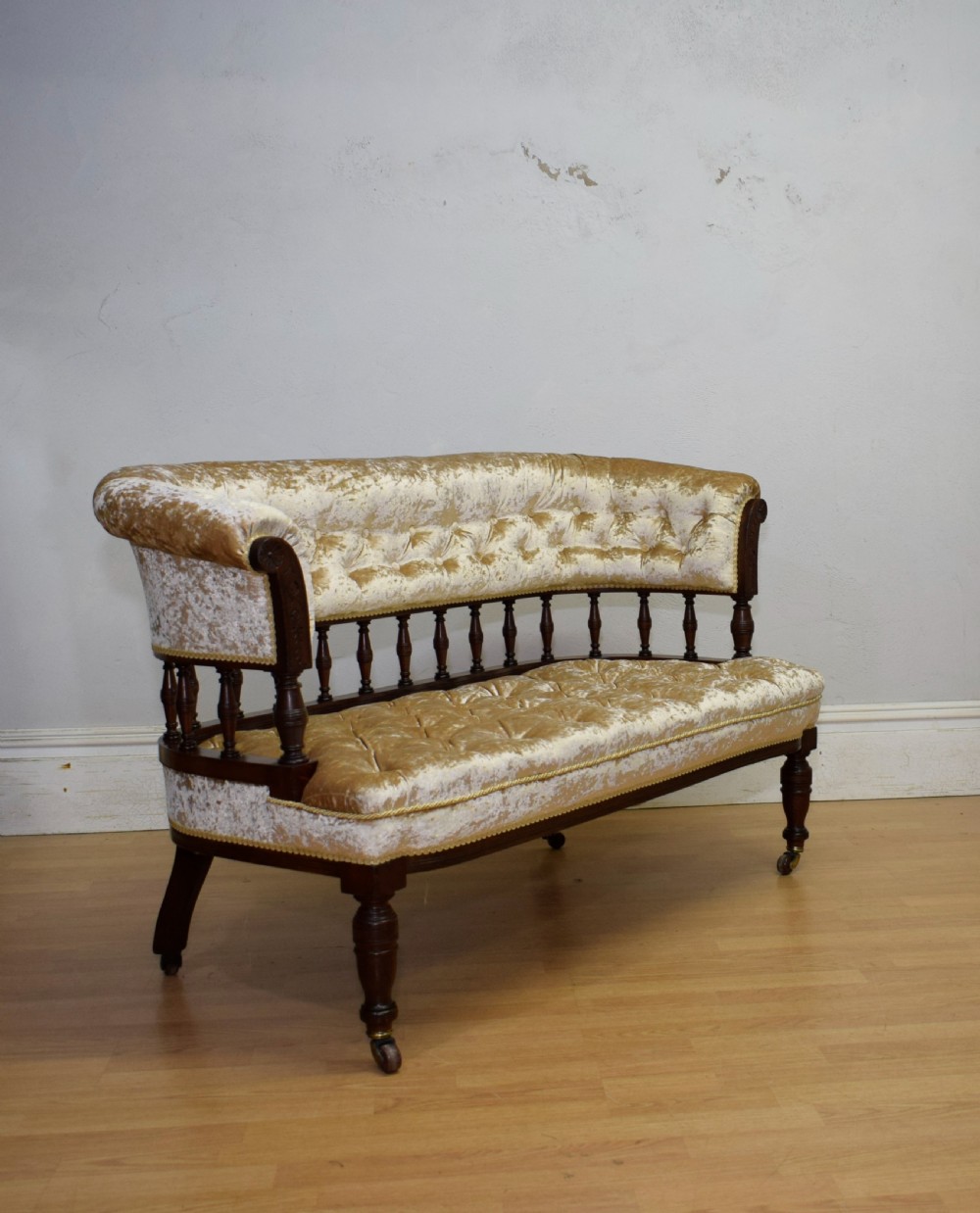 A Victorian mahogany settee with champagne gold crushed velvet deep-buttoned upholstery, a spindle gallery back, and turned legs with castors. View V