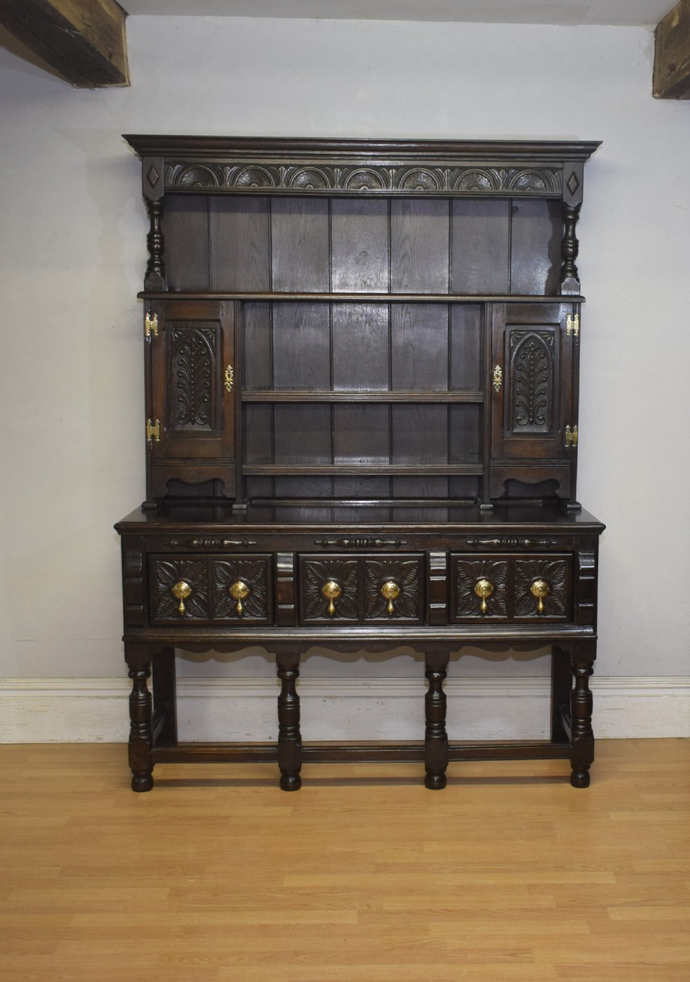 A dark carved oak Jacobean style Welsh dresser featuring an upper plate rack with side cupboard doors over a lower sideboard base with three carved drawers and turned legs. View II