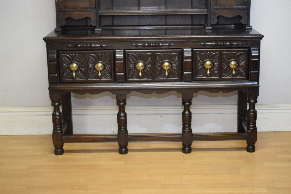 A dark carved oak Jacobean style Welsh dresser featuring an upper plate rack with side cupboard doors over a lower sideboard base with three carved drawers and turned legs. View V