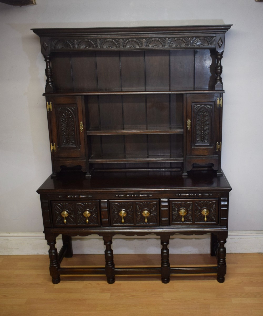 A dark carved oak Jacobean style Welsh dresser featuring an upper plate rack with side cupboard doors over a lower sideboard base with three carved drawers and turned legs. View VI