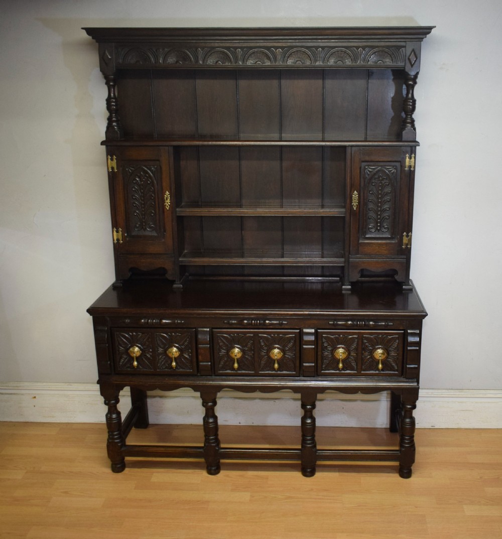 A dark carved oak Jacobean style Welsh dresser featuring an upper plate rack with side cupboard doors over a lower sideboard base with three carved drawers and turned legs. View VII