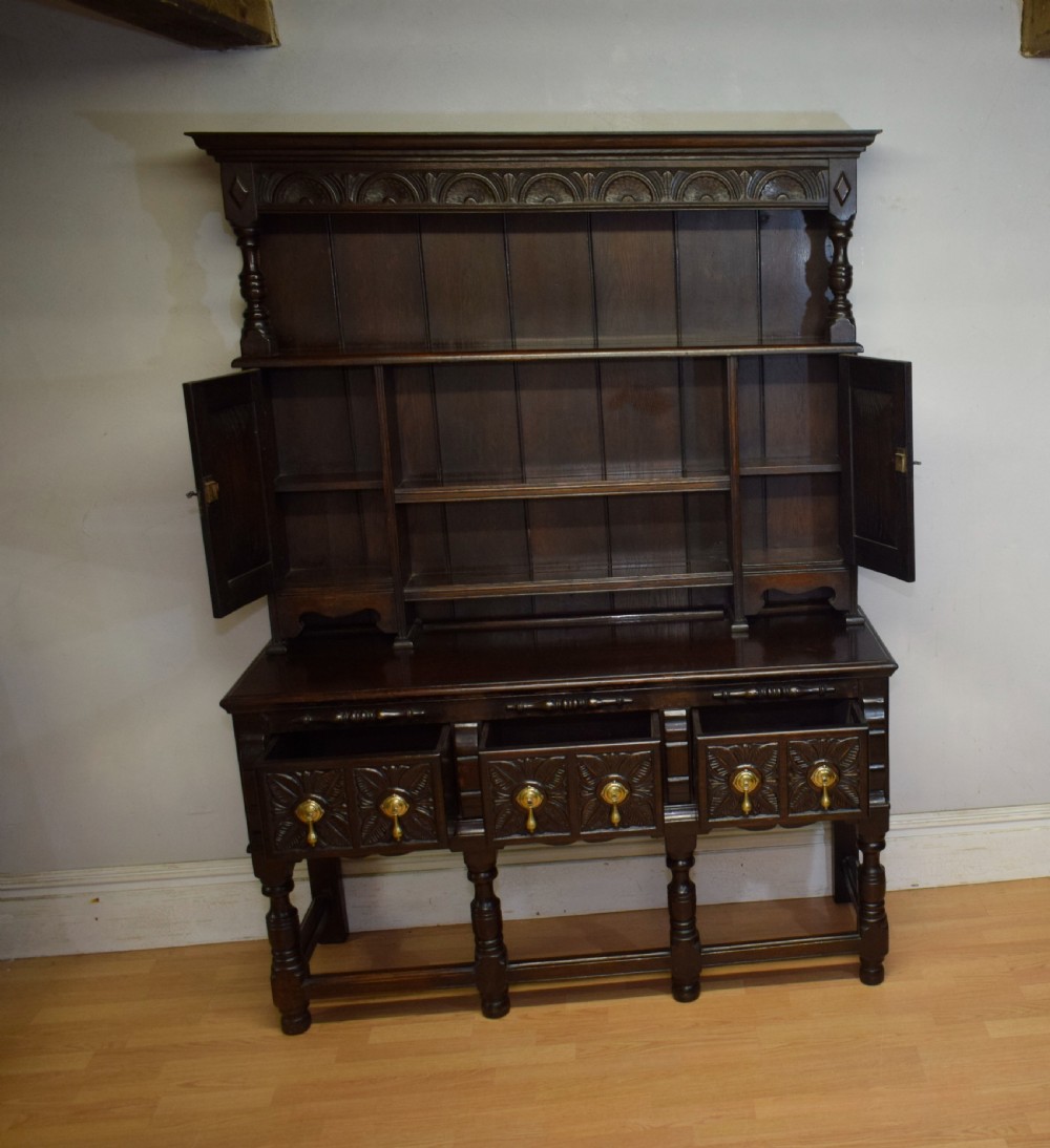 A dark carved oak Jacobean style Welsh dresser featuring an upper plate rack with side cupboard doors over a lower sideboard base with three carved drawers and turned legs. View XVII