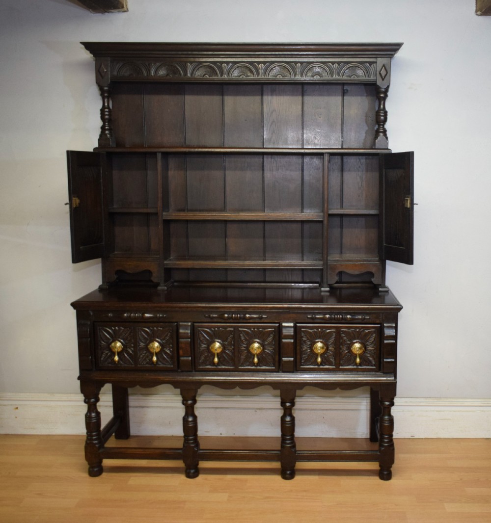 A dark carved oak Jacobean style Welsh dresser featuring an upper plate rack with side cupboard doors over a lower sideboard base with three carved drawers and turned legs. View XVIII