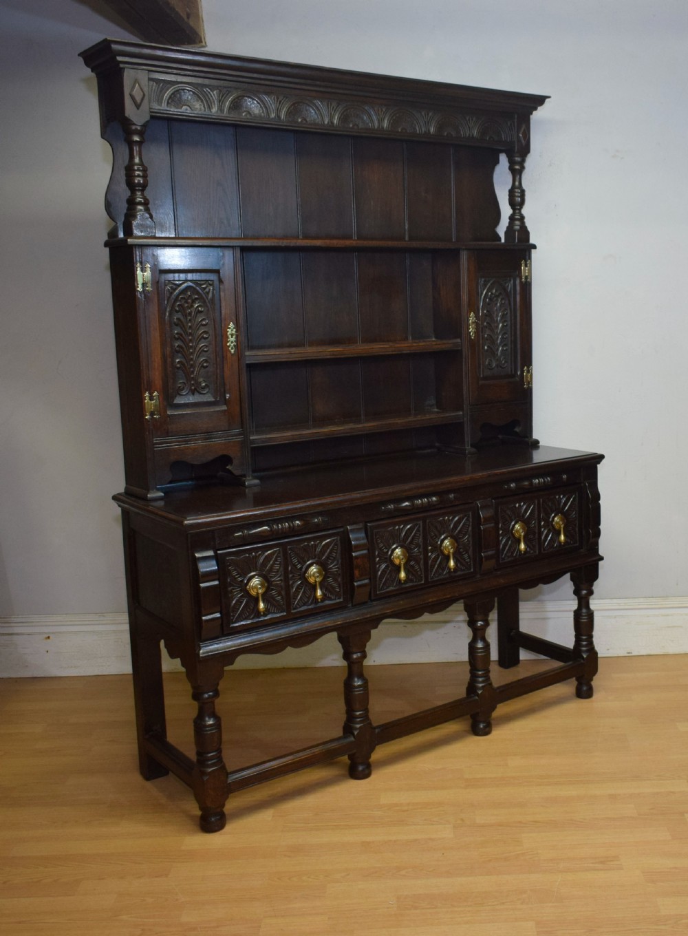 A dark carved oak Jacobean style Welsh dresser featuring an upper plate rack with side cupboard doors over a lower sideboard base with three carved drawers and turned legs. View XIX