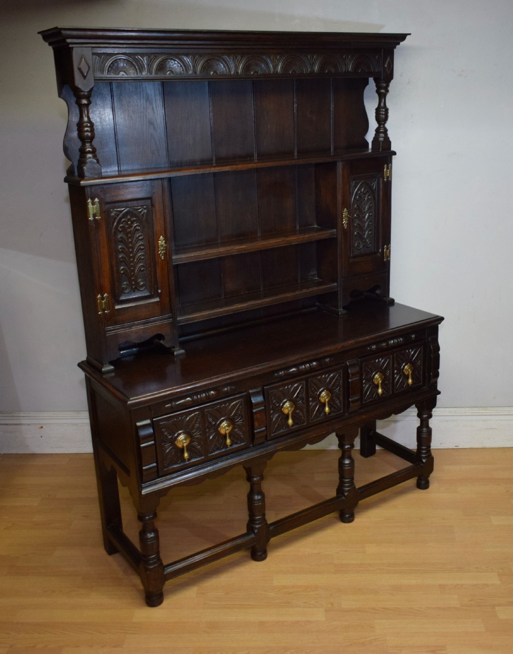 A dark carved oak Jacobean style Welsh dresser featuring an upper plate rack with side cupboard doors over a lower sideboard base with three carved drawers and turned legs. View XX