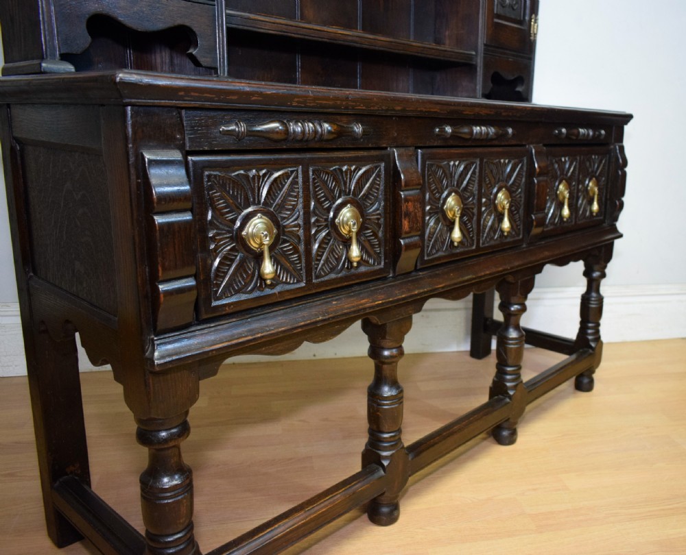 A dark carved oak Jacobean style Welsh dresser featuring an upper plate rack with side cupboard doors over a lower sideboard base with three carved drawers and turned legs. View XXI