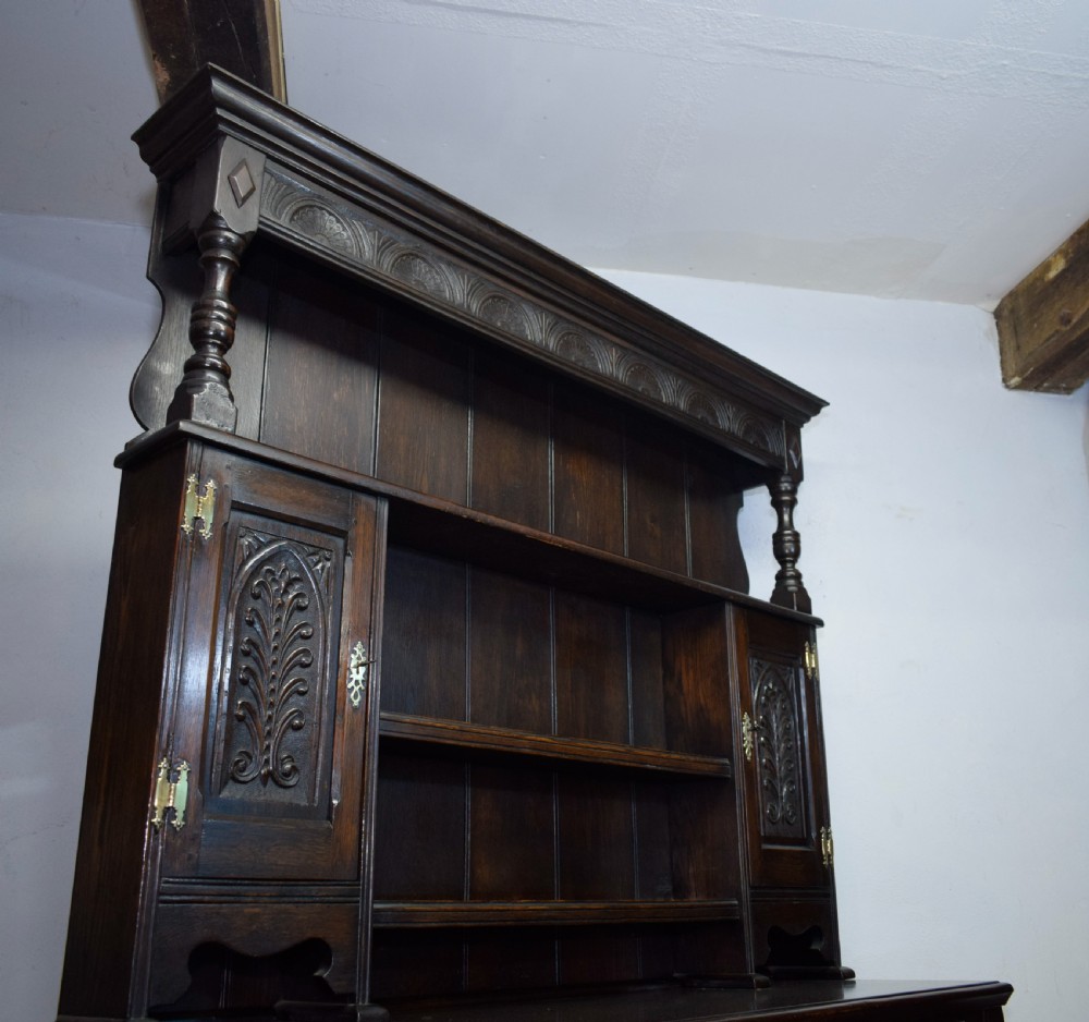 A dark carved oak Jacobean style Welsh dresser featuring an upper plate rack with side cupboard doors over a lower sideboard base with three carved drawers and turned legs. View XXII
