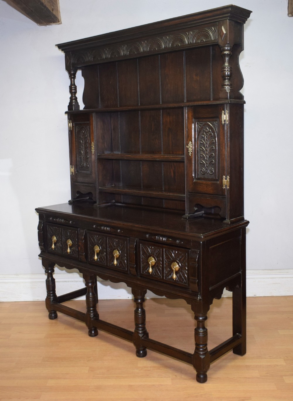 A dark carved oak Jacobean style Welsh dresser featuring an upper plate rack with side cupboard doors over a lower sideboard base with three carved drawers and turned legs. View XXIII