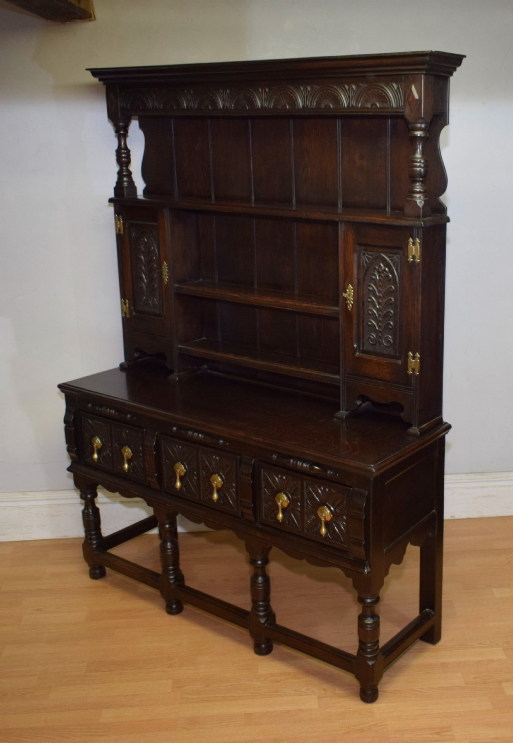 A dark carved oak Jacobean style Welsh dresser featuring an upper plate rack with side cupboard doors over a lower sideboard base with three carved drawers and turned legs. View XXIV