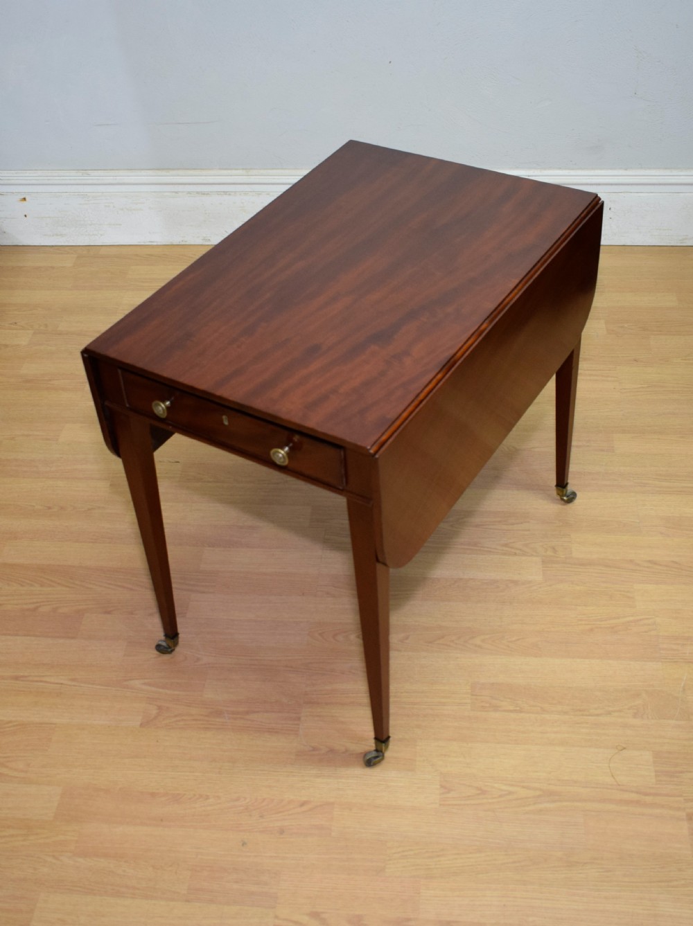 A Regency mahogany Pembroke table featuring dual drop-leaf sides, a front drawer with two brass pull knobs, square tapered legs, and brass brass castors.
