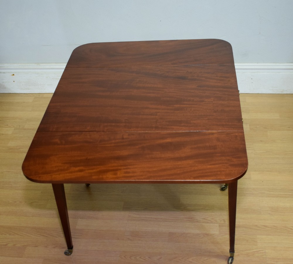 A Regency mahogany Pembroke table featuring dual drop-leaf sides, a front drawer with two brass pull knobs, square tapered legs, and brass brass castors. View XXI