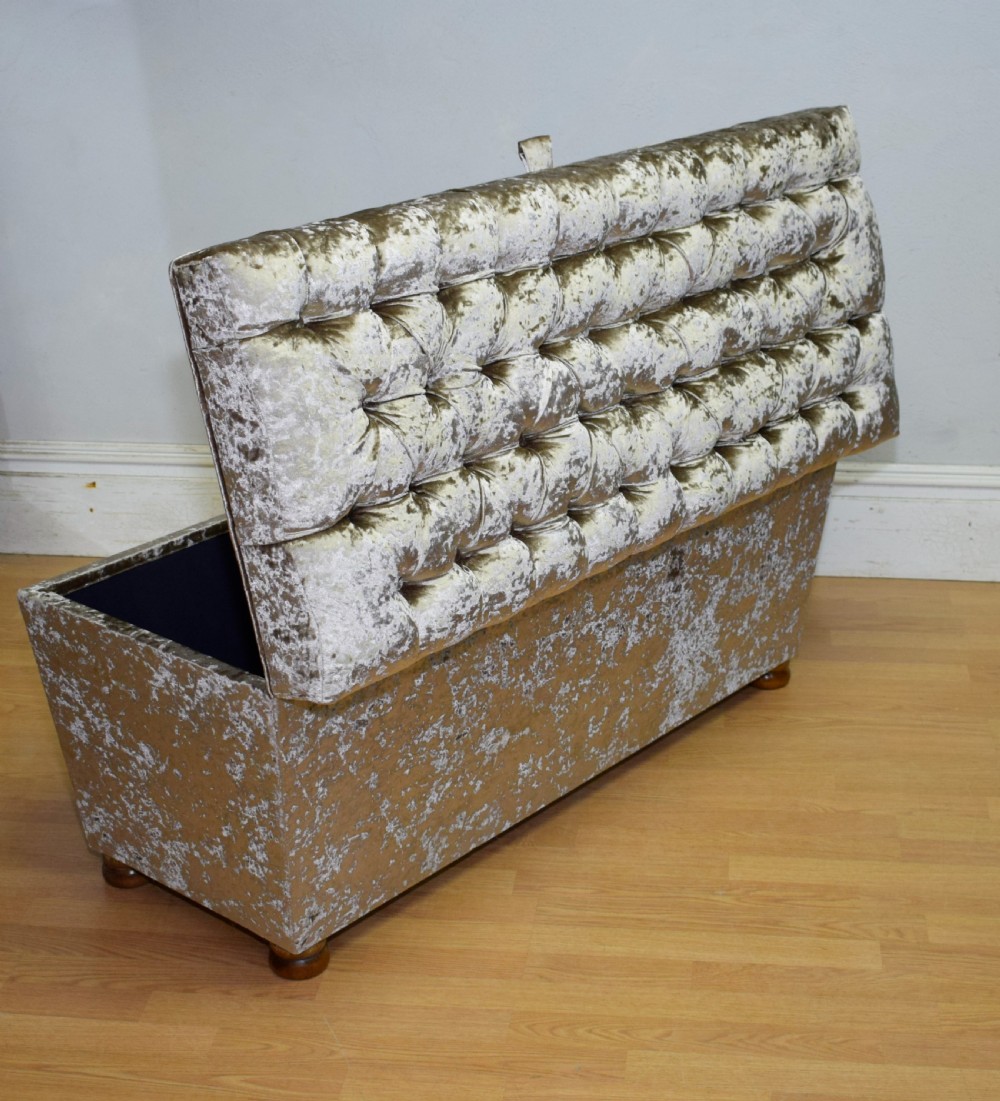 A rectangular upholstered storage ottoman in champagne crushed velvet with a deep buttoned tufted lid and small wooden feet. View VI