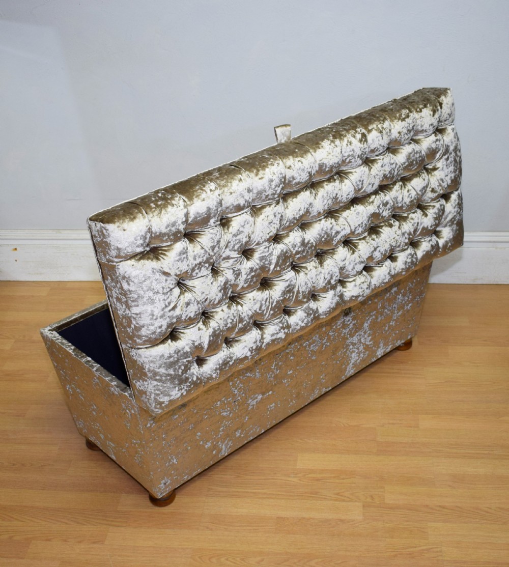 A rectangular upholstered storage ottoman in champagne crushed velvet with a deep buttoned tufted lid and small wooden feet. View VIII