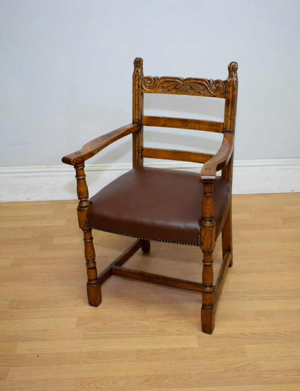 A carved oak armchair featuring a brown leather seat with stud detailing, turned front legs, and carved crest rail on the backrest.