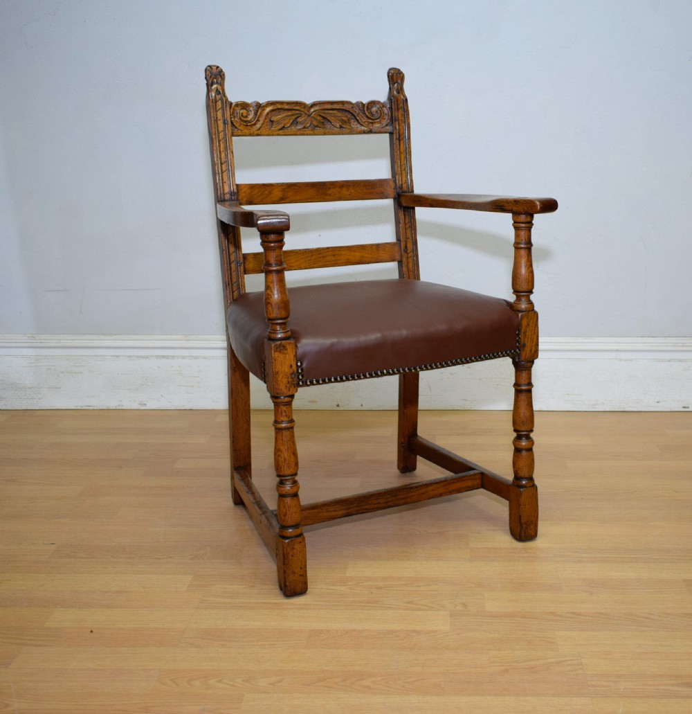 A carved oak armchair featuring a brown leather seat with stud detailing, turned front legs, and carved crest rail on the backrest. View V