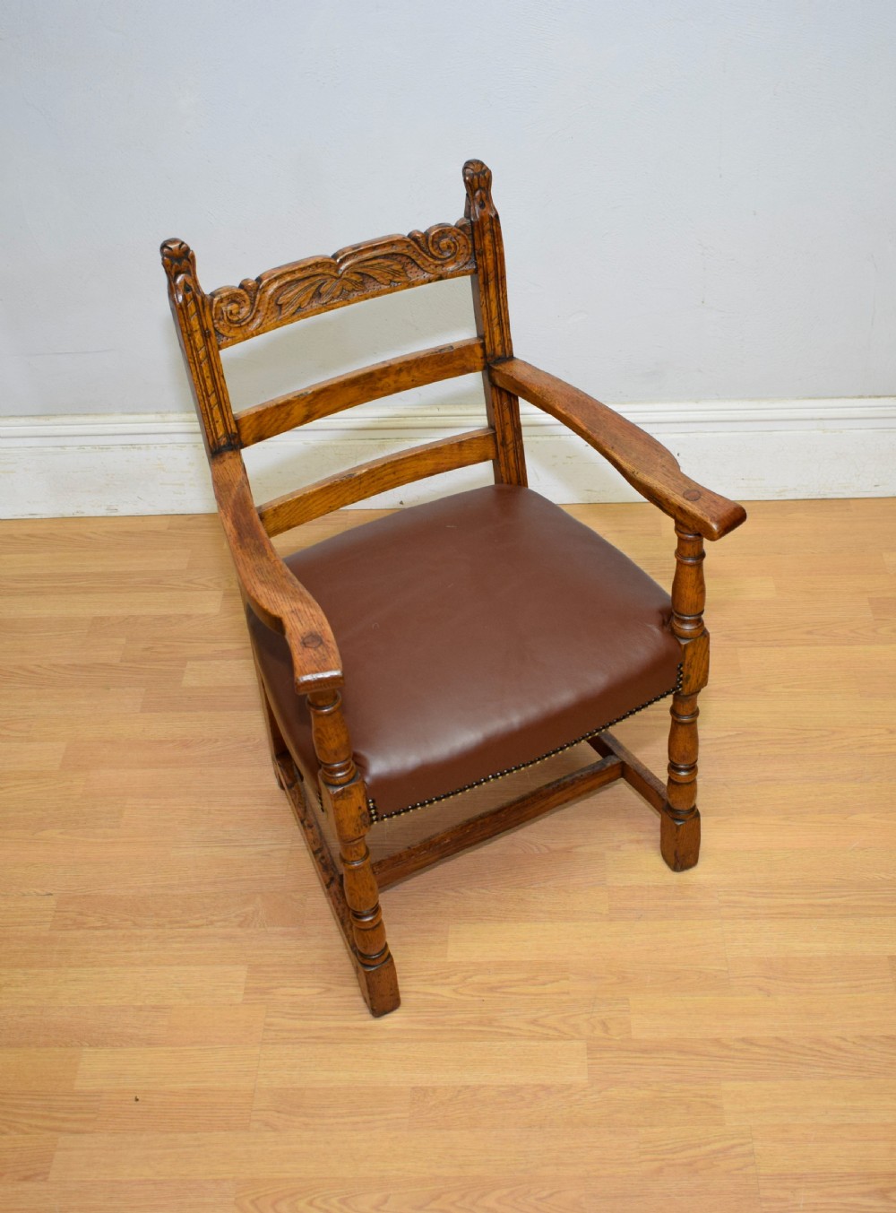 A carved oak armchair featuring a brown leather seat with stud detailing, turned front legs, and carved crest rail on the backrest. View VII