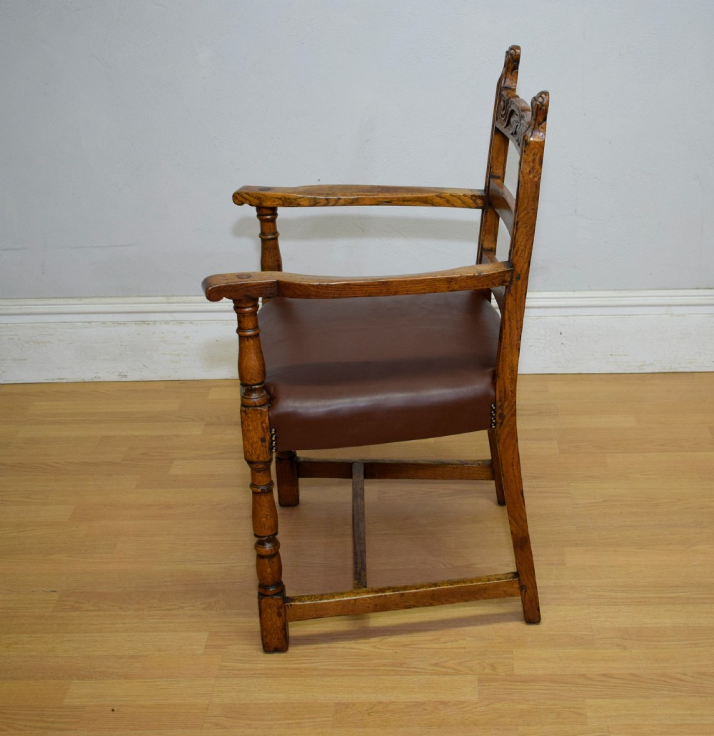 A carved oak armchair featuring a brown leather seat with stud detailing, turned front legs, and carved crest rail on the backrest. View XV