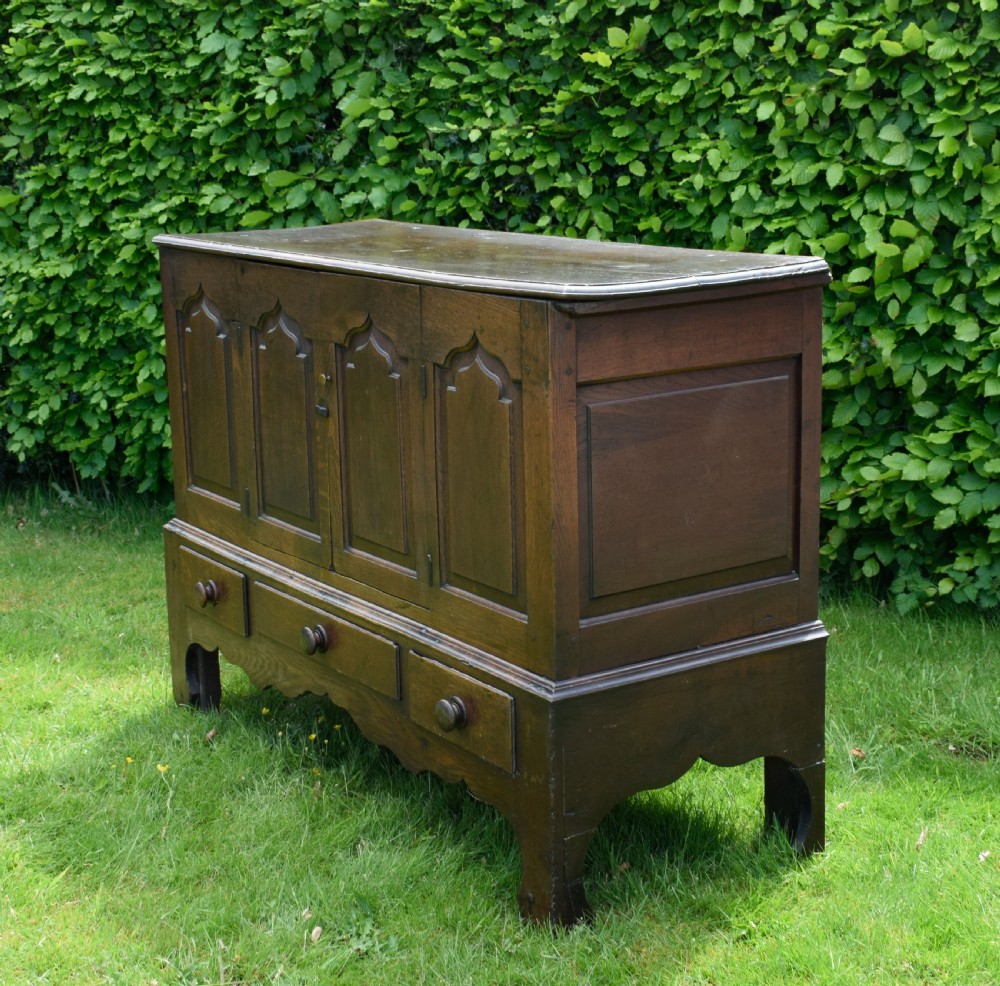 A 17th-century style dark oak court cupboard with four arched fielded panel doors over three drawers fitted with turned wooden knobs. View V