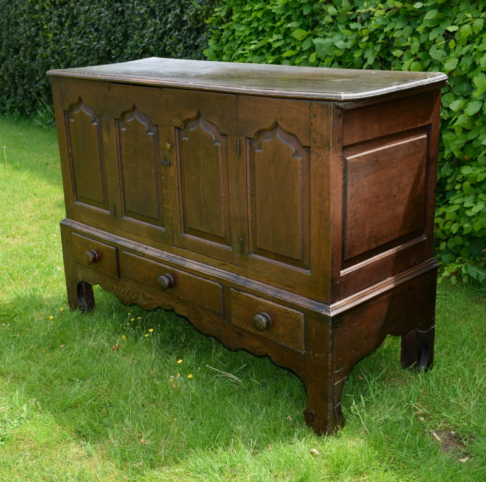A 17th-century style dark oak court cupboard with four arched fielded panel doors over three drawers fitted with turned wooden knobs. View VI