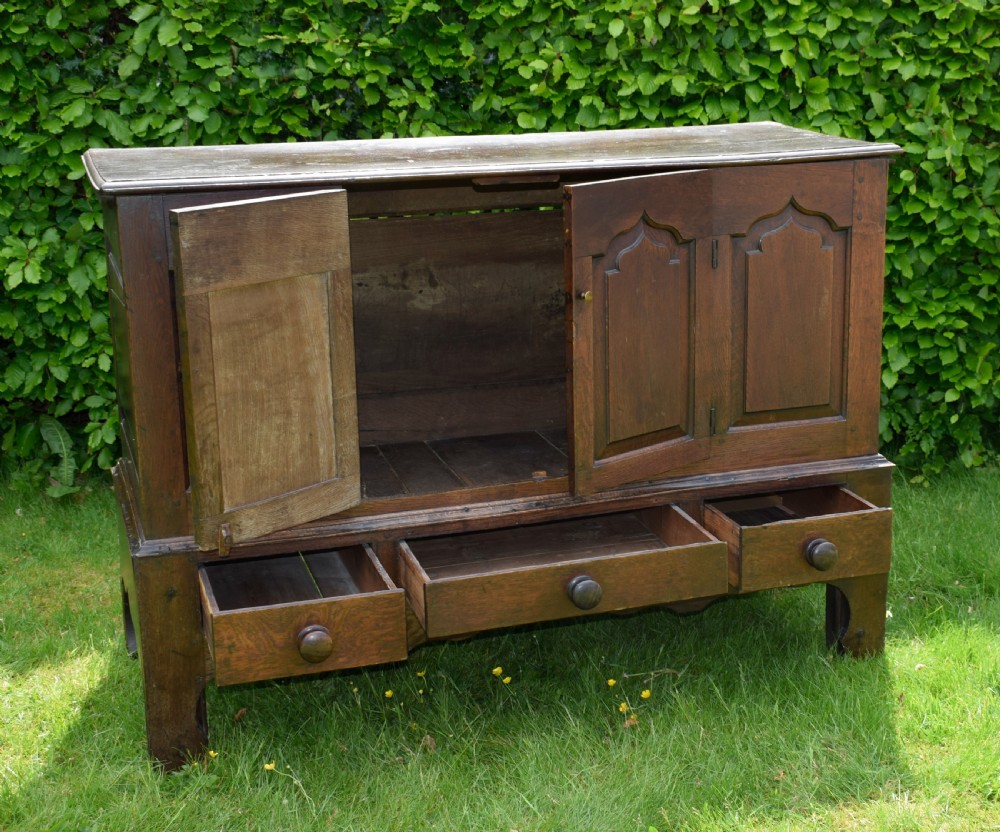 A 17th-century style dark oak court cupboard with four arched fielded panel doors over three drawers fitted with turned wooden knobs. View XII
