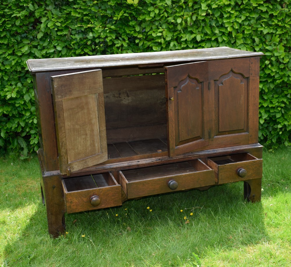 A 17th-century style dark oak court cupboard with four arched fielded panel doors over three drawers fitted with turned wooden knobs. View XIV