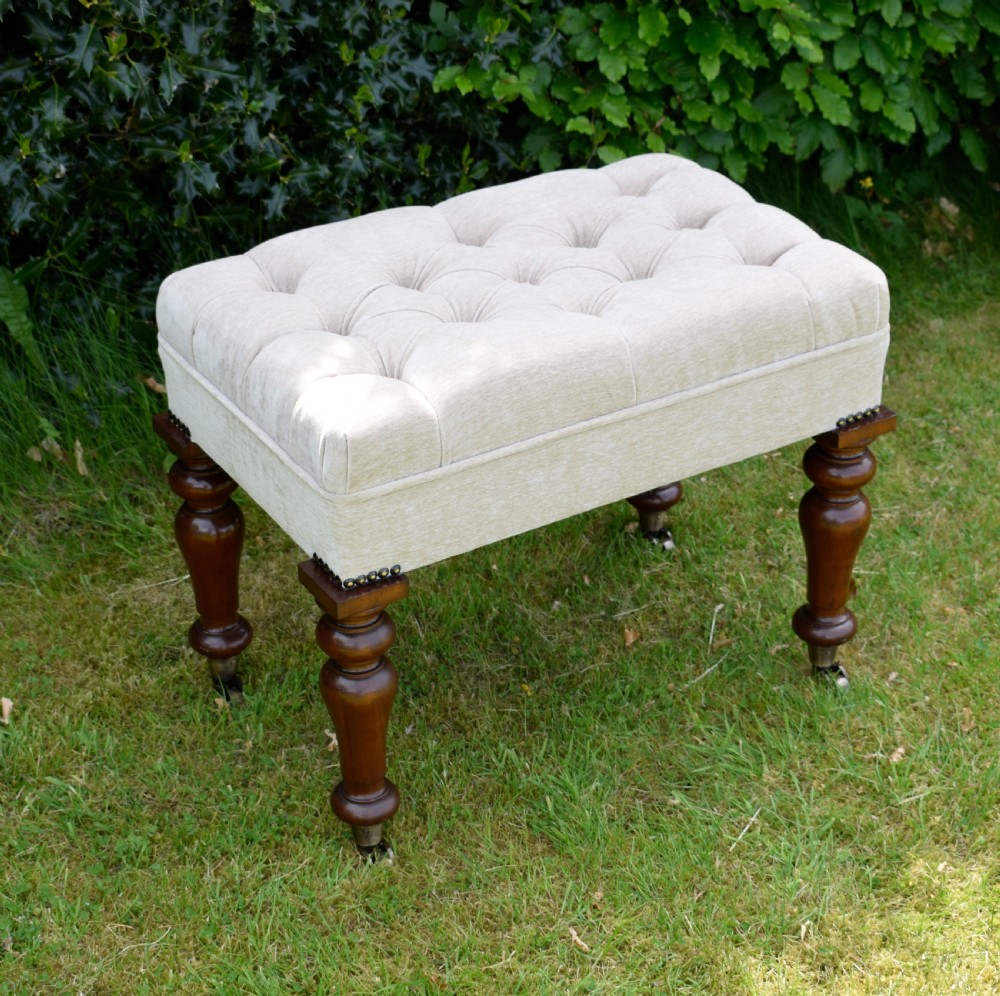 A rectangular cream fabric upholstered stool featuring deep button tufting, brass nailhead trim, and dark turned wooden legs with brass castors.