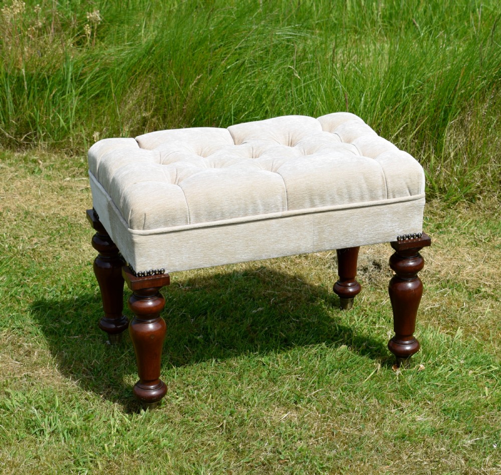 A rectangular cream fabric upholstered stool featuring deep button tufting, brass nailhead trim, and dark turned wooden legs with brass castors. View V