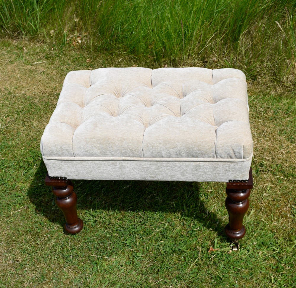 A rectangular cream fabric upholstered stool featuring deep button tufting, brass nailhead trim, and dark turned wooden legs with brass castors. View VI