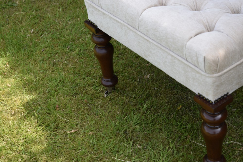 A rectangular cream fabric upholstered stool featuring deep button tufting, brass nailhead trim, and dark turned wooden legs with brass castors. View X
