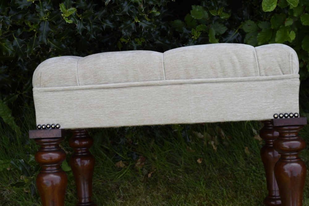 A rectangular cream fabric upholstered stool featuring deep button tufting, brass nailhead trim, and dark turned wooden legs with brass castors. View XVII