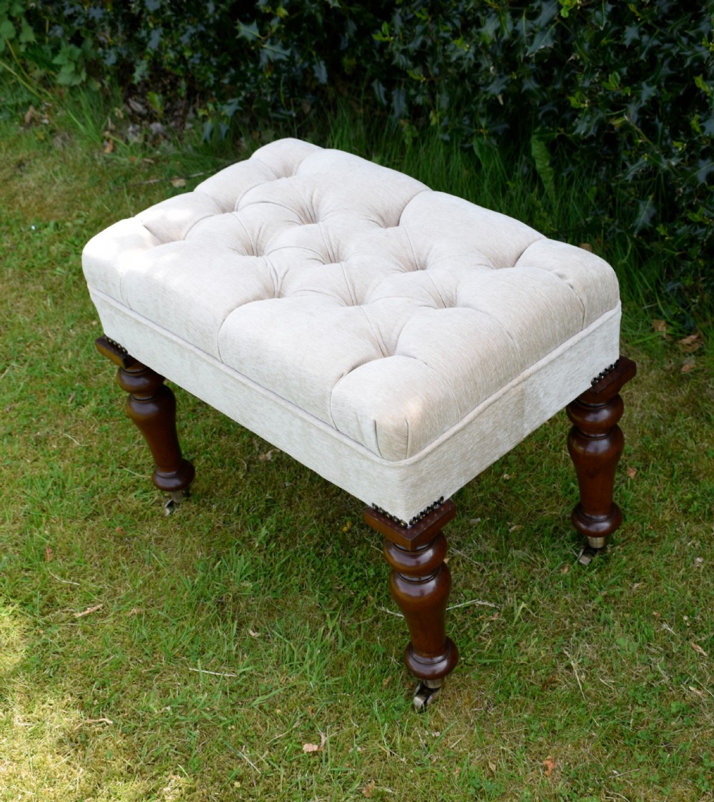 A rectangular cream fabric upholstered stool featuring deep button tufting, brass nailhead trim, and dark turned wooden legs with brass castors. View XXI