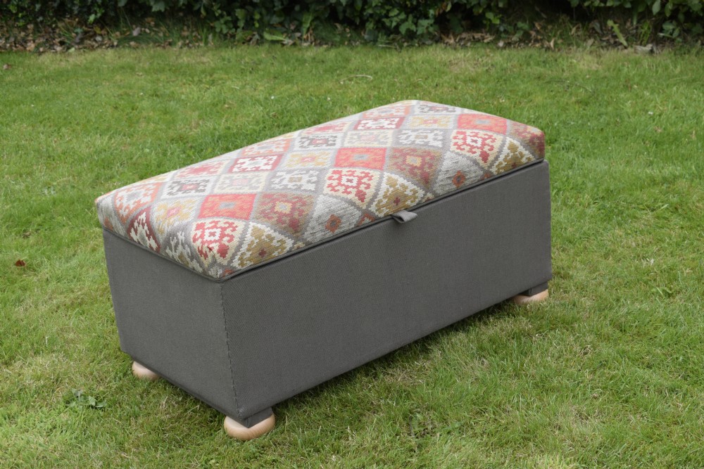 A rectangular upholstered storage ottoman featuring a dark grey base, short turned wooden feet, and a geometric kilim-patterned hinged top.