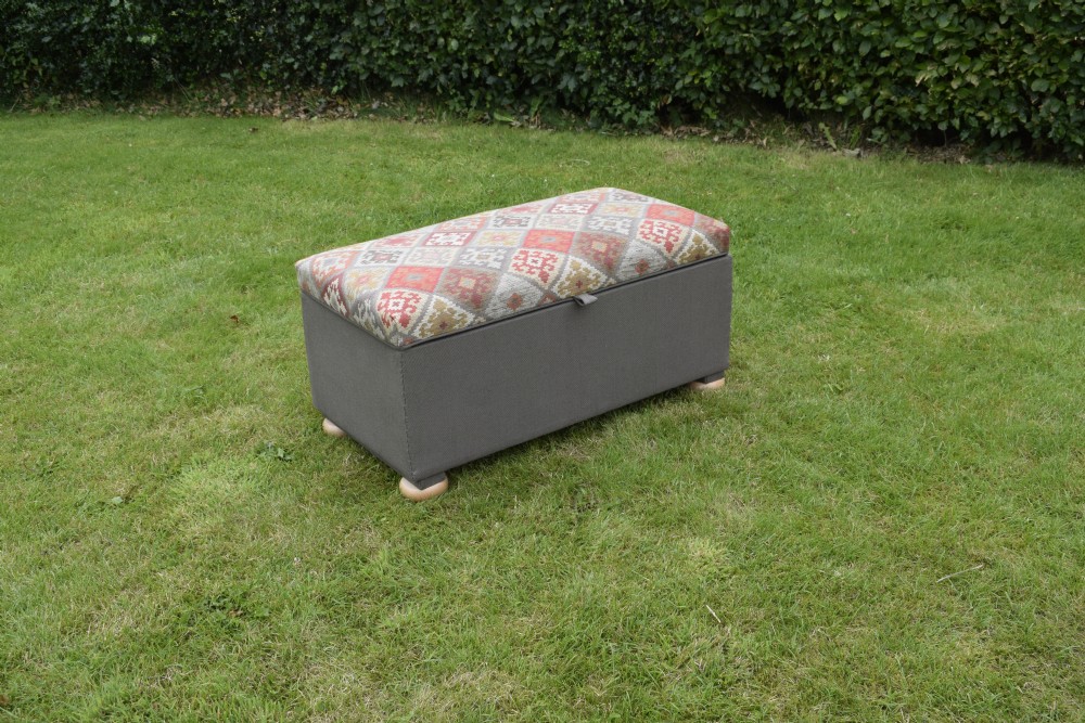 A rectangular upholstered storage ottoman featuring a dark grey base, short turned wooden feet, and a geometric kilim-patterned hinged top. View III