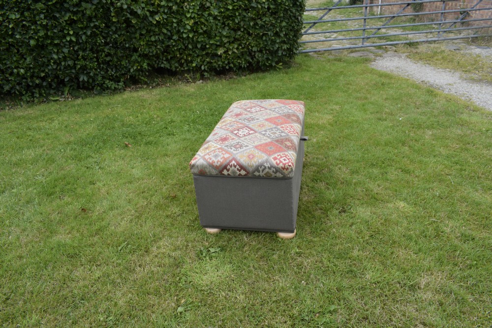 A rectangular upholstered storage ottoman featuring a dark grey base, short turned wooden feet, and a geometric kilim-patterned hinged top. View V