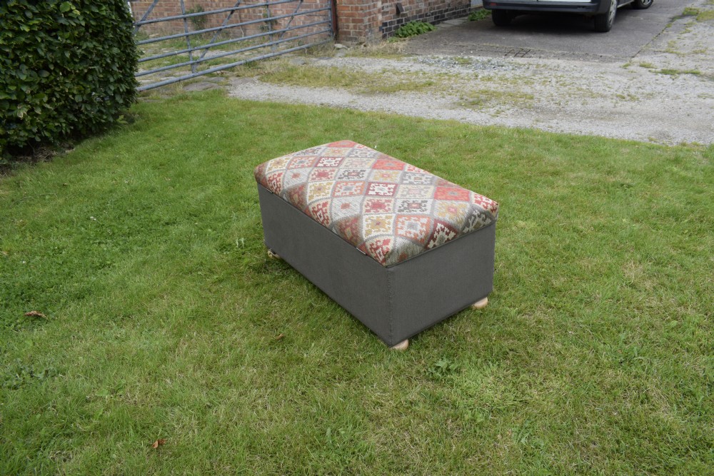 A rectangular upholstered storage ottoman featuring a dark grey base, short turned wooden feet, and a geometric kilim-patterned hinged top. View VI