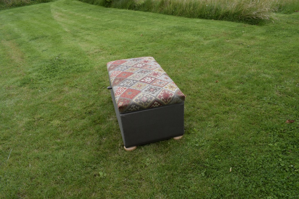 A rectangular upholstered storage ottoman featuring a dark grey base, short turned wooden feet, and a geometric kilim-patterned hinged top. View VII