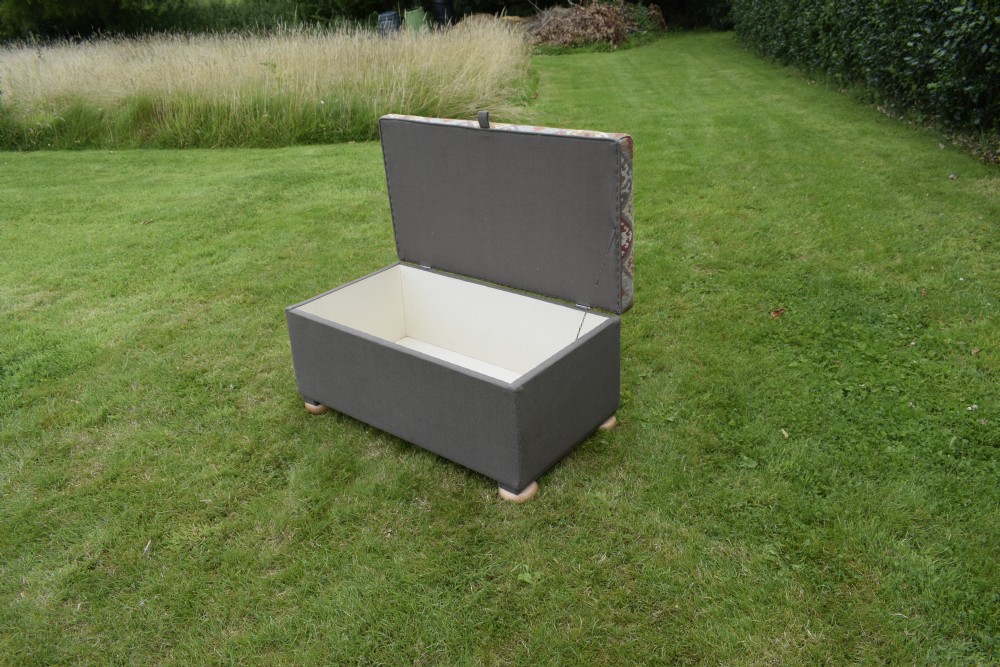 A rectangular upholstered storage ottoman featuring a dark grey base, short turned wooden feet, and a geometric kilim-patterned hinged top. View IX