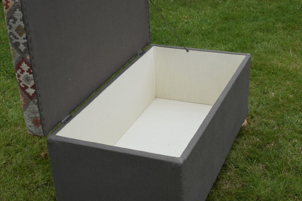 A rectangular upholstered storage ottoman featuring a dark grey base, short turned wooden feet, and a geometric kilim-patterned hinged top. View X