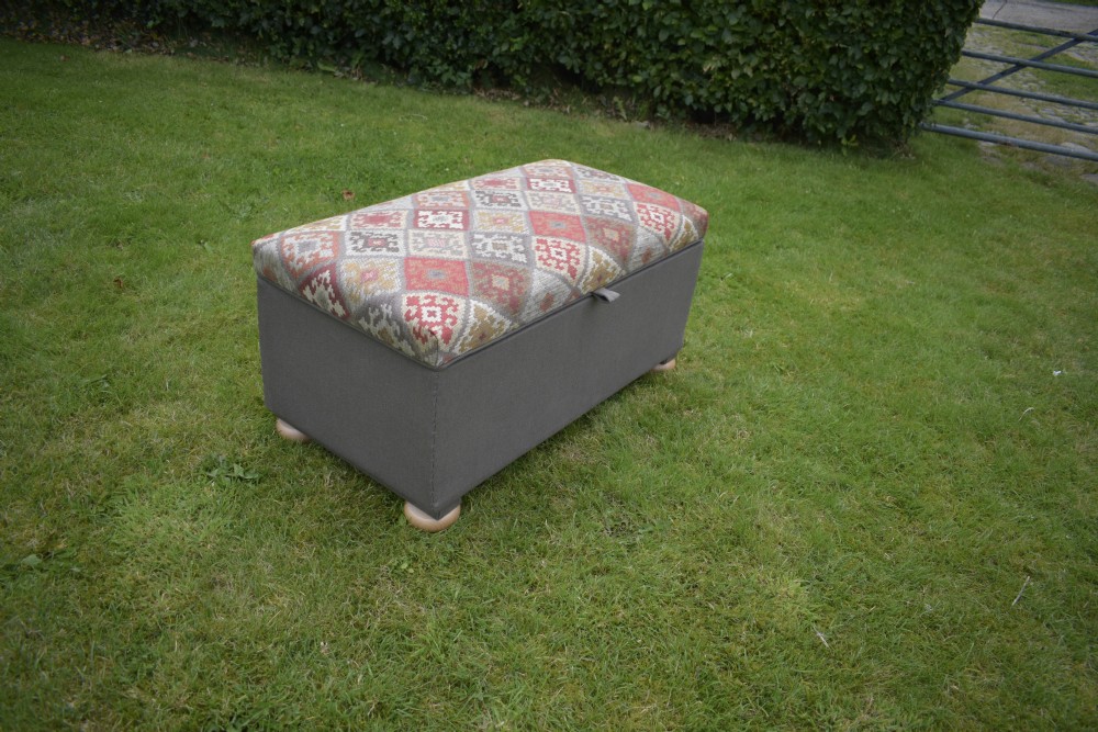 A rectangular upholstered storage ottoman featuring a dark grey base, short turned wooden feet, and a geometric kilim-patterned hinged top. View XI