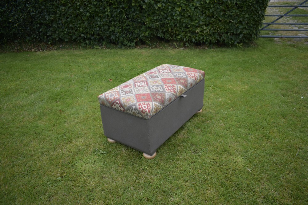 A rectangular upholstered storage ottoman featuring a dark grey base, short turned wooden feet, and a geometric kilim-patterned hinged top. View XII