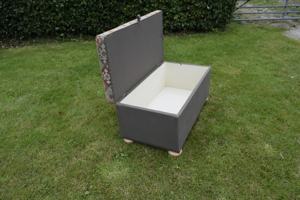 A rectangular upholstered storage ottoman featuring a dark grey base, short turned wooden feet, and a geometric kilim-patterned hinged top. View XIII