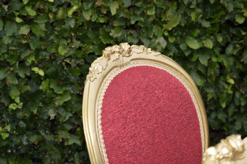 A set of four French-style dining chairs featuring gold-painted frames with carved oval backs and red woven fabric upholstery. View X