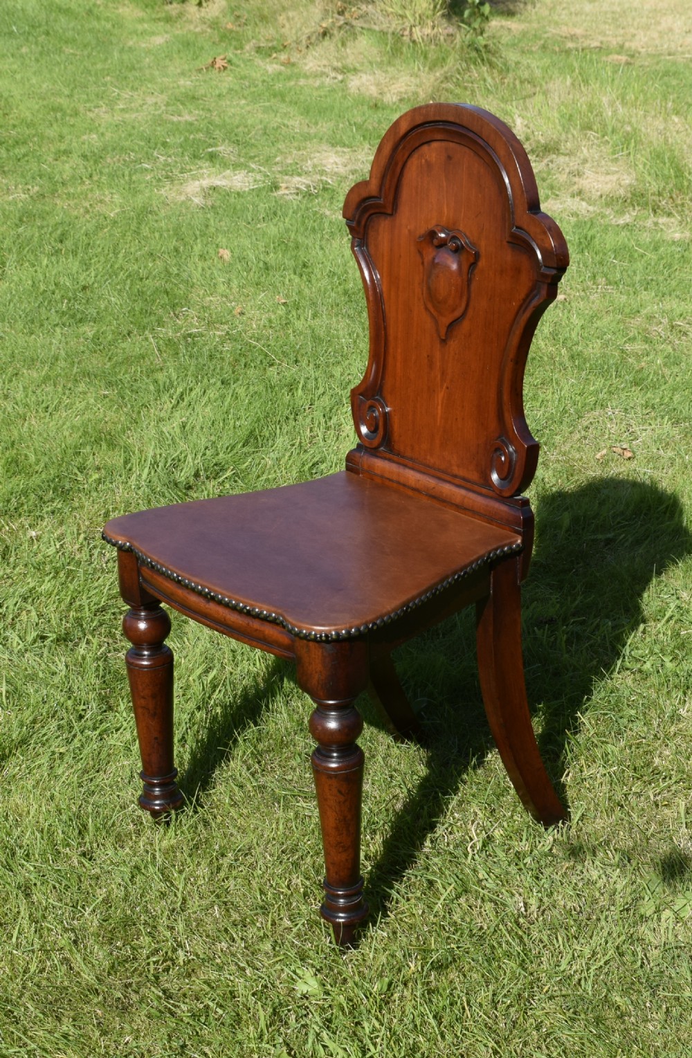 A Victorian mahogany hall chair with a carved shield-shaped backrest, stud-trimmed dark seat, turned front legs, and splayed rear legs. View V
