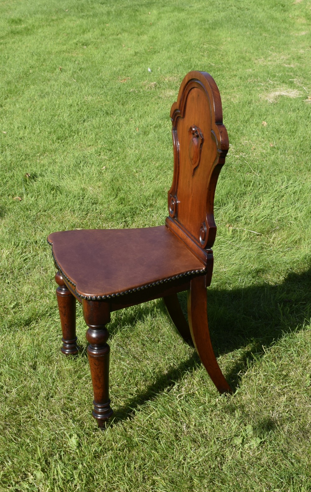 A Victorian mahogany hall chair with a carved shield-shaped backrest, stud-trimmed dark seat, turned front legs, and splayed rear legs. View VI