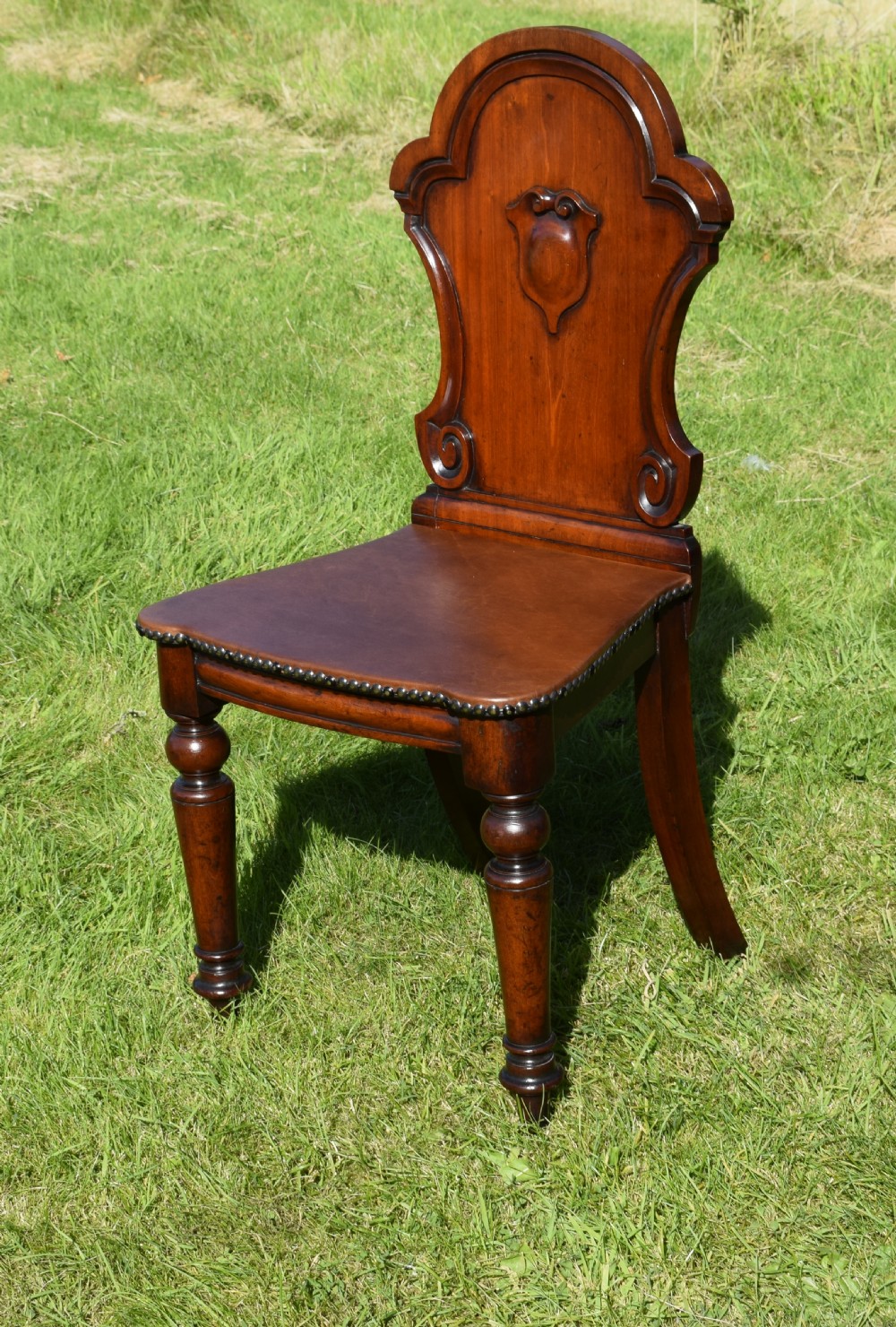 A Victorian mahogany hall chair with a carved shield-shaped backrest, stud-trimmed dark seat, turned front legs, and splayed rear legs. View VIII