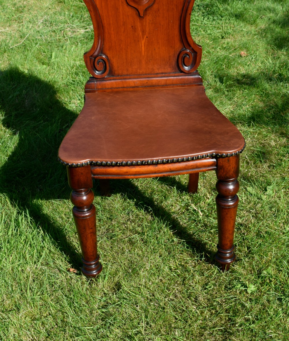 A Victorian mahogany hall chair with a carved shield-shaped backrest, stud-trimmed dark seat, turned front legs, and splayed rear legs. View X
