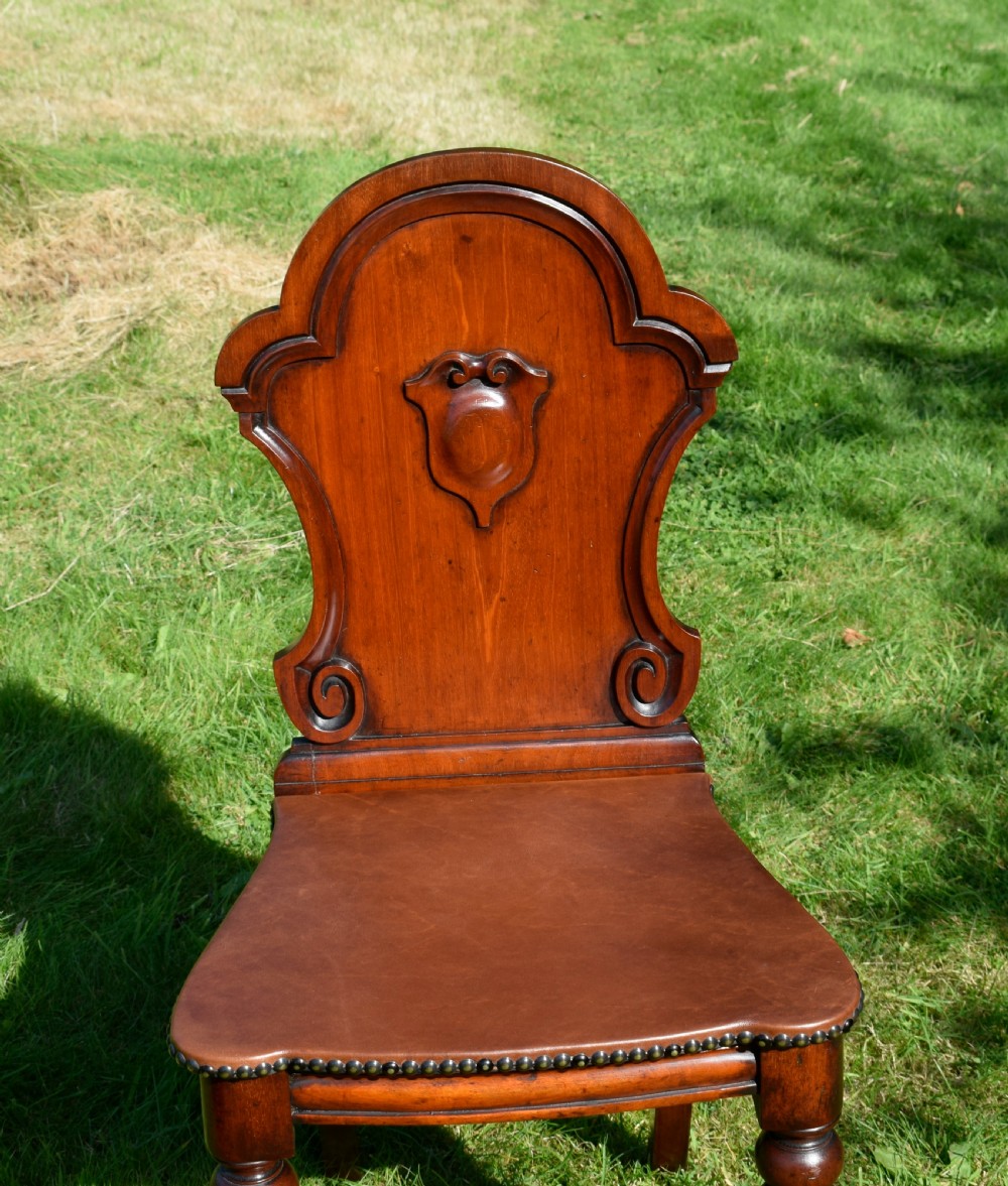 A Victorian mahogany hall chair with a carved shield-shaped backrest, stud-trimmed dark seat, turned front legs, and splayed rear legs. View XI